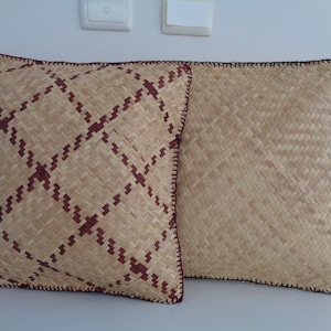 May include: Two woven cushions with a brown and beige pattern. The cushion on the left has a more intricate pattern with a red accent. Both cushions have a brown crocheted border.