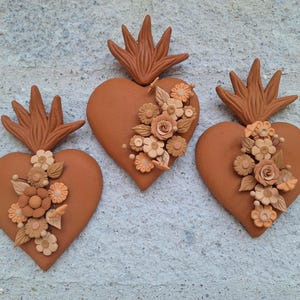 May include: Three terracotta-colored heart-shaped wall decorations. Each heart is adorned with a floral arrangement in shades of beige and orange. A stylized flame design sits atop each heart.