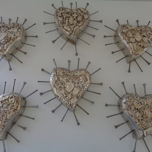 Puede incluir: Cinco adornos de pared en forma de coraz&oacute;n de tono plateado dispuestos en grupo. Cada coraz&oacute;n presenta una superficie texturizada con varias figuras peque&ntilde;as en relieve y est&aacute; rodeado de clavos met&aacute;licos que irradian desde los bordes.