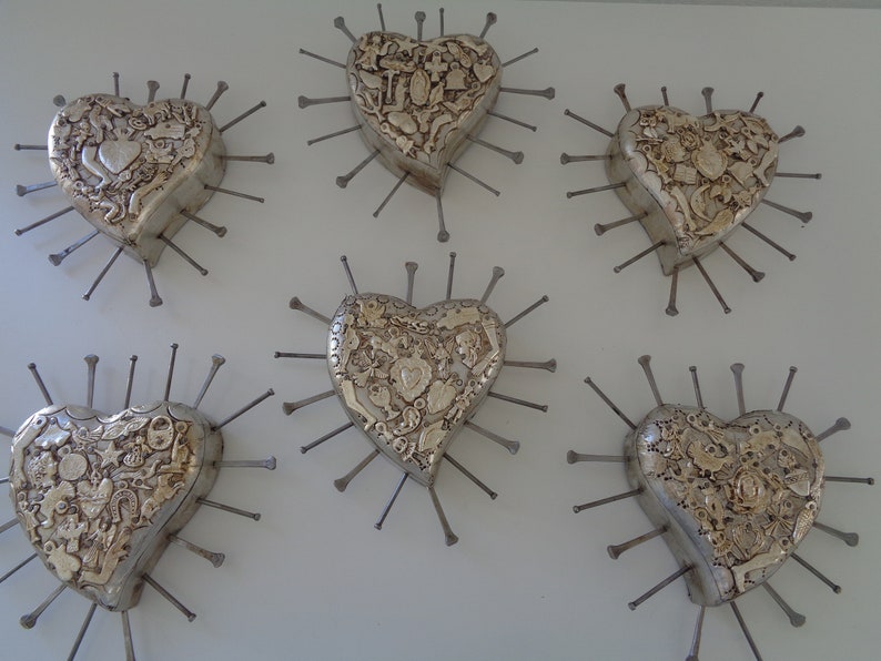 Puede incluir: Cinco adornos de pared en forma de coraz&oacute;n de tono plateado dispuestos en grupo. Cada coraz&oacute;n presenta una superficie texturizada con varias figuras peque&ntilde;as en relieve y est&aacute; rodeado de clavos met&aacute;licos que irradian desde los bordes.