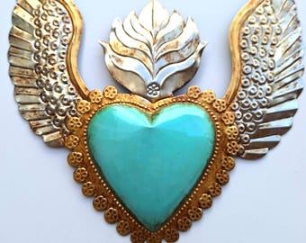 Puede incluir: Un corazón decorativo de metal con esmalte turquesa, ribete dorado y alas plateadas. El corazón está rodeado por un borde dorado con un borde festoneado. Un elemento floral plateado se encuentra encima del corazón.