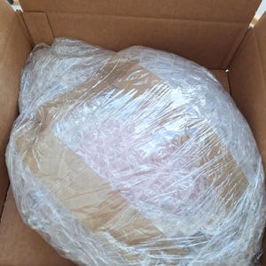 Puede incluir: Una caja de cart&oacute;n marr&oacute;n que contiene un art&iacute;culo envuelto. El art&iacute;culo est&aacute; protegido por film transparente y pl&aacute;stico de burbujas. El contenido es parcialmente visible a trav&eacute;s del embalaje.