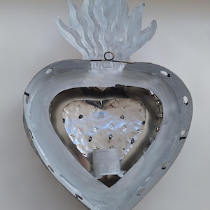 Puede incluir: Decoraci&oacute;n de pared met&aacute;lica en forma de coraz&oacute;n con dise&ntilde;o de llama en la parte superior. Pintada en tonos grises y plateados, con interior texturizado y un portavelas. Ideal para decoraci&oacute;n de interiores.