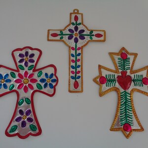 May include: Three silver metal crosses with colorful floral and heart designs. The crosses are decorated with red, pink, green, blue, and purple paint.