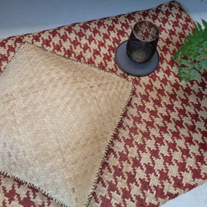 May include: A woven, light brown, square cushion with black stitching sits on a red and white woven rug. A black metal candle holder sits on a black coaster on the rug.