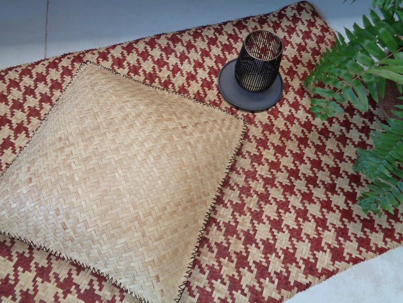 May include: A woven, light brown, square cushion with black stitching sits on a red and white woven rug. A black metal candle holder sits on a black coaster on the rug.