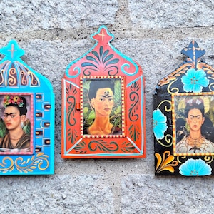 May include: Three decorative wall-mounted frames featuring portraits of Frida Kahlo. The left frame is bright blue with gold accents, the center frame is orange with red and green details, and the right frame is black with blue floral motifs.