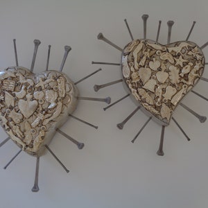 Puede incluir: Dos decoraciones de pared en forma de coraz&oacute;n con una superficie texturizada de color blanco roto cubierta por varias formas peque&ntilde;as moldeadas. Cada coraz&oacute;n est&aacute; rodeado por largos y finos clavos met&aacute;licos oscuros que irradian hacia afuera.
