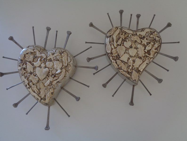 Puede incluir: Dos decoraciones de pared en forma de coraz&oacute;n con una superficie texturizada de color blanco roto cubierta por varias formas peque&ntilde;as moldeadas. Cada coraz&oacute;n est&aacute; rodeado por largos y finos clavos met&aacute;licos oscuros que irradian hacia afuera.