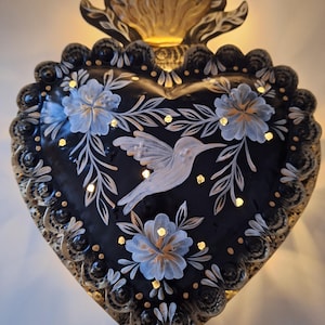 Puede incluir: Decoraci&oacute;n de pared en forma de coraz&oacute;n negro con un dise&ntilde;o de colibr&iacute; y flores. La pieza de arte met&aacute;lica presenta una base negra con detalles pintados en blanco y azul, y un borde dorado. La parte superior tiene un dise&ntilde;o de llama dorada, y peque&ntilde;as luces incrustadas.