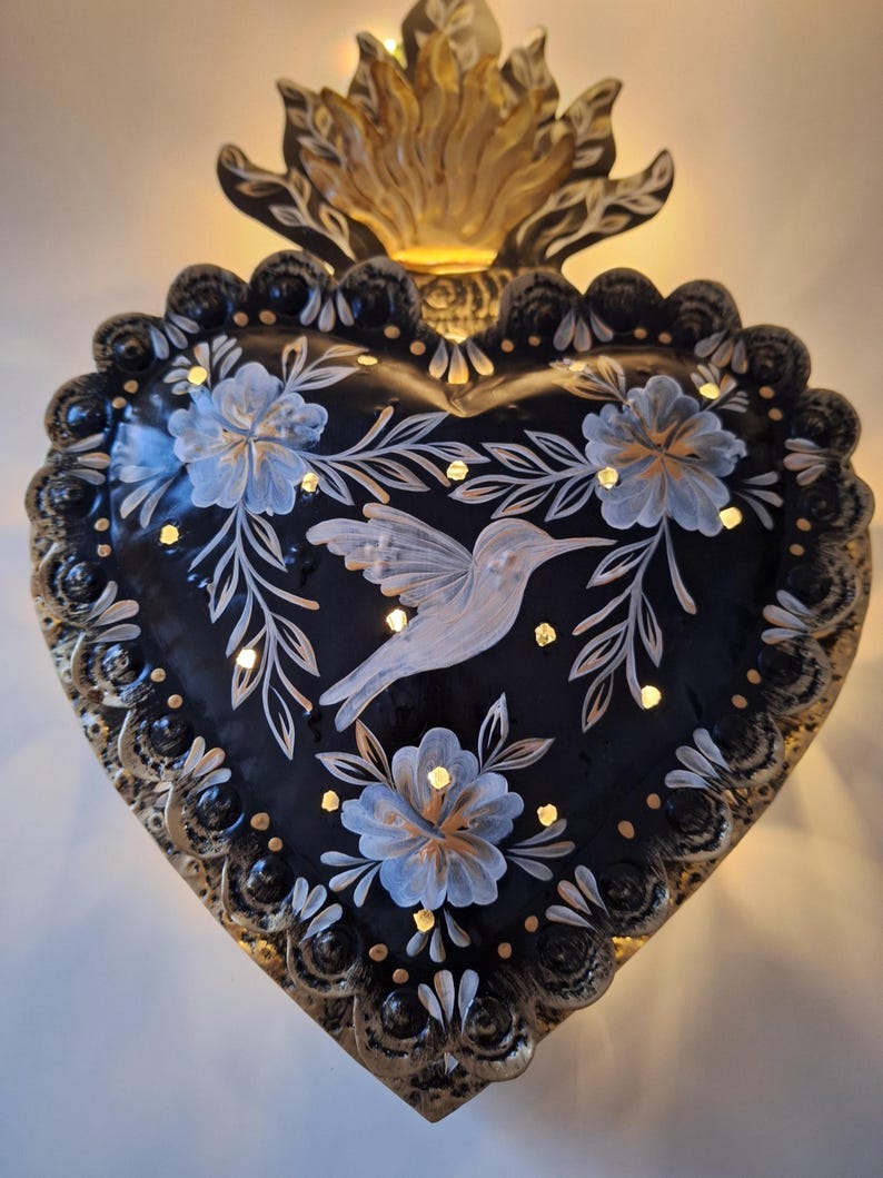 Puede incluir: Decoraci&oacute;n de pared en forma de coraz&oacute;n negro con un dise&ntilde;o de colibr&iacute; y flores. La pieza de arte met&aacute;lica presenta una base negra con detalles pintados en blanco y azul, y un borde dorado. La parte superior tiene un dise&ntilde;o de llama dorada, y peque&ntilde;as luces incrustadas.