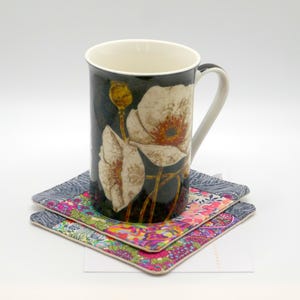 May include: A tall ceramic mug with a floral design featuring large white and orange flowers on a dark background. The mug sits on two square coasters with colorful floral patterns. The handle is white.