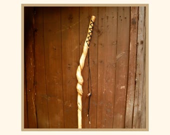 May include: A tall, light-colored wooden walking stick featuring a natural, twisted grain pattern along its length. The upper section is wrapped with black cord in a crisscross pattern, and a long black cord hangs down with a small wooden bead.