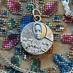 Puede incluir: Un collar con colgante de plata con un medallón circular que representa a una mujer con un tocado. El medallón lleva la inscripción "B.E. TAVERNER CAMELIN" y "1872". El collar está sobre un fondo de cuentas de colores.