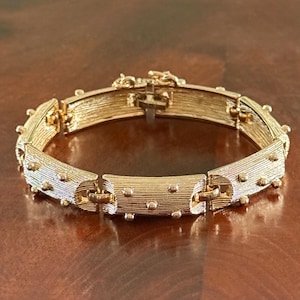Könnte beinhalten: Ein goldfarbenes Armband mit einem strukturierten, geriffelten Design. Das Armband hat kleine, gleichmäßig verteilte, runde Akzente und einen sicheren Verschluss. Das Schmuckstück wird auf einer dunkelbraunen Oberfläche präsentiert.