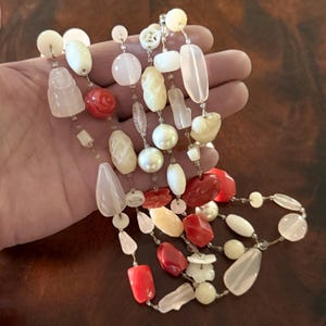 May include: A multi-strand necklace featuring a variety of beads in shades of white, pink, and red. The beads include carved shapes, pearls, and irregular stones, strung on a delicate chain. The necklace is displayed against a neutral background.