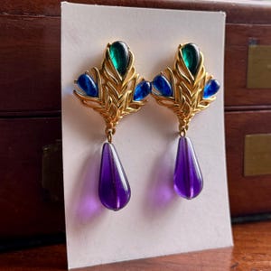 May include: A pair of gold-tone earrings featuring a fan-shaped design with inset green and blue teardrop-shaped stones. Each earring is finished with a dangling, translucent purple teardrop-shaped pendant suspended from the base of the metal frame.