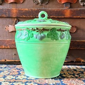Peut inclure: Un pot en céramique vert vif avec un couvercle assorti présentant un motif en relief de vignes brunes, de feuilles vertes et de grappes de raisin. Le couvercle possède une poignée circulaire au sommet et le pot a un corps lisse et conique.