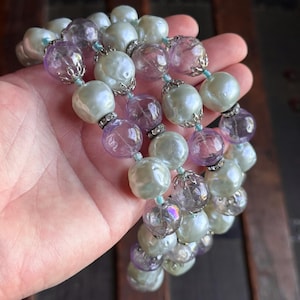 Könnte beinhalten: Eine Vintage-Halskette mit großen, irisierenden Kunstperlen, die mit klaren und lavendelfarbenen Glasperlen durchsetzt sind. Die Perlen sind mit silberfarbenen Metallakzenten und kleinen blauen Perlen verbunden. Die Halskette wird in einer Hand gehalten.