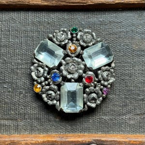 Könnte beinhalten: Eine silberfarbene Vintage-Brosche mit floralem Design. Die Brosche hat drei große, rechteckige, hellblaue Edelsteine und mehrere kleinere, farbige Edelsteine. Die Brosche ist vor einem dunkelgrauen Hintergrund platziert.