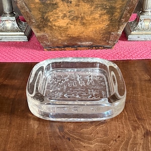 Peut inclure: Un cendrier carré en verre transparent aux coins arrondis. Le cendrier présente un motif gravé de figures et de symboles. Le cendrier est posé sur une surface en bois.