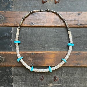 May include: A white shell necklace with turquoise accents and silver beads. The necklace is laid out on a wooden surface.