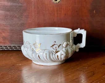 Puede incluir: Taza de afeitar de cerámica blanca con un diseño floral dorado y un asa. La taza tiene una parte superior dividida para contener jabón de afeitar y un cepillo.