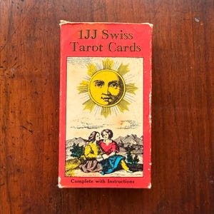 Puede incluir: Una caja rectangular para 1JJ Swiss Tarot Cards con un borde rojo. El frente presenta un sol amarillo con un rostro humano sobre una ilustración de dos personas sentadas en un paisaje. El texto dice 1JJ Swiss Tarot Cards y Complete with Instructions.