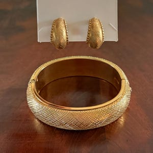 Könnte beinhalten: Ein goldfarbenes Schmuckset mit einem strukturierten Armreif und passenden Creolen. Das Armband hat einen Klappverschluss und ein gewebtes Muster. Die Ohrringe sind kleine, strukturierte Creolen mit einem Perlenrand.