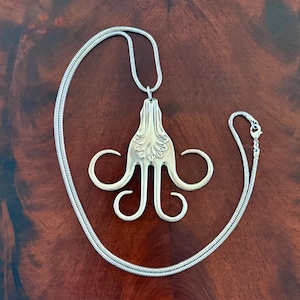 May include: A silver pendant necklace with a distinctive design. The pendant, resembling an octopus, is made from a repurposed fork. The necklace has a snake chain and a lobster clasp. The pendant is approximately 7.6 cm wide.