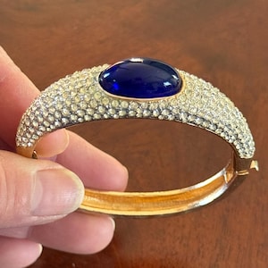 May include: A gold-toned hinged bracelet with a large, oval, deep blue stone at the center. The bracelet is encrusted with small, clear crystals. The bracelet is held open by a hand, revealing the gold interior.