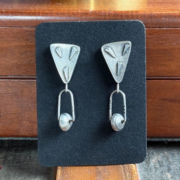 May include: A pair of silver-toned earrings displayed on a black card. Each earring features a triangular top with cut-out shapes, connected to a curved wire and a small, round bead. The earrings are presented against a wooden background.
