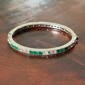 Puede incluir: Brazalete rígido plateado con bisagras y piedras cuadradas alternas de color verde esmeralda y transparente. El brazalete tiene un diseño grabado alrededor de los bordes y está sobre una superficie de madera marrón oscuro.