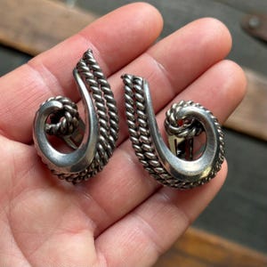 May include: A pair of silver-toned, vintage earrings. Each earring features a swirling, open design with a rope-like texture. The earrings are held in a hand, showcasing their intricate details and classic style. The earrings have a clip-on style.