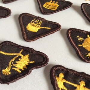 May include: A collection of triangular embroidered patches featuring dark brown fabric with gold thread designs. Each patch displays a unique icon, including a birthday cake, a steaming pot, a pine tree, a globe with stars, and stylised figures.