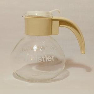 May include: A clear glass carafe with a cream-coloured handle and lid. The word "Whistler" is printed on the glass. The handle is curved and attached to the carafe's body. The lid has a small spout.