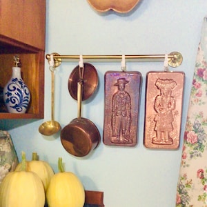 May include: A brass wall-mounted rack holds a brass ladle, a small copper saucepan with a lid, and two rectangular copper moulds featuring embossed figures of a person in a hat and a person with a headpiece, all hanging from white hooks.