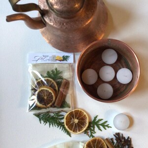 May include: A hammered copper kettle, a copper bowl containing five white tea light candles, and a clear plastic bag labelled Simmer Pot with dried orange slices, cinnamon sticks, and evergreen sprigs. Loose dried orange slices, cloves, and a candle are included.