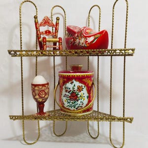 May include: A gold-coloured metal shelf unit with two tiers, displaying decorative items. A miniature red chair and a red clog with floral designs are on the top shelf. The bottom shelf holds an egg cup with an egg, and a red and white floral container.