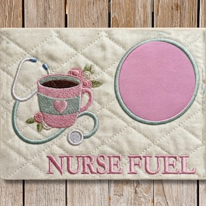 以下が含まれることがあります： ピンクとティール色のコーヒーカップのデザイン、聴診器、ピンクの円が刺繍されたクリーム色のマット。「NURSE FUEL」の文字がピンク色で刺繍されています。マットはキルティング模様です。
