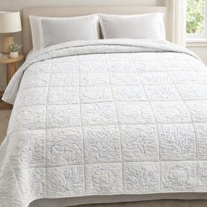 May include: A white quilt featuring a grid pattern with stitched outlines of various sea life, including starfish, coral, and seashells. The quilt is draped over a bed with two matching white pillows, providing a clean and textured appearance.