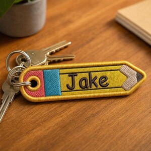 May include: A yellow pencil-shaped embroidered keychain featuring a pink eraser, a blue band, and the name Jake stitched in black thread. The keychain includes a metal ring attached to the eraser end, with silver-colored keys connected to it.