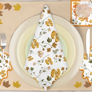 May include: A fall-themed table setting with a white napkin with a floral pattern, two forks, a knife, a spoon, and two leaf-shaped placemats with a floral pattern. The placemats are orange and brown with a white floral pattern.