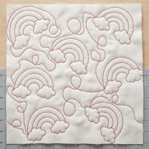 May include: A square, off-white fabric panel featuring a repeating pattern of embroidered rainbows and fluffy clouds in a reddish-brown thread. The design includes various sizes of rainbows and clouds, creating a whimsical and cheerful aesthetic. The panel is placed on a wooden surface.