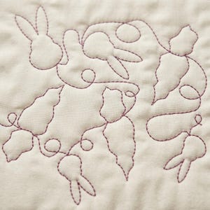 May include: Close-up of a quilted fabric featuring a repeating pattern of rabbits and carrots. The design is stitched in a dark purple thread on a cream-colored background. The rabbits have long ears and the carrots have pointed ends.