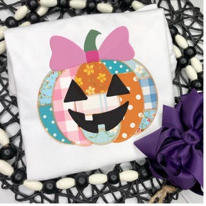 May include: White t-shirt featuring a patchwork pumpkin design with a pink bow. The pumpkin has a smiling face and is made of various patterned fabrics in orange, blue, pink, and yellow. A purple bow is also visible.