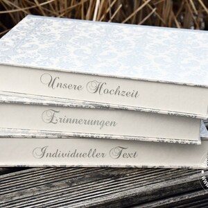 Könnte beinhalten: Drei gestapelte dekorative Boxen mit einem floralen Muster auf den Deckeln. Die Boxen sind cremefarben mit grauem Text, der "Unsere Hochzeit", "Erinnerungen" und "Individueller Text" lautet. Die Boxen stehen auf einer Holzoberfläche.