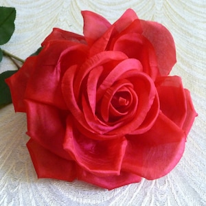 May include: A single, large, red silk rose with a green stem and leaves. The rose is arranged on a white, textured surface.