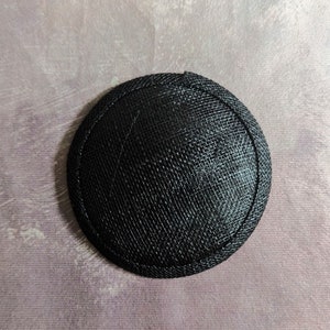 May include: A black round hat base made of woven material. The base is about 4 inches in diameter.