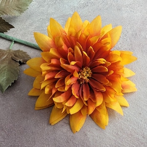 May include: A single, large, orange and yellow artificial flower with a layered, ruffled texture. The flower is on a gray background.